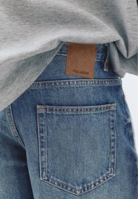Gros plan sur la poche arrière d'un jean en denim bleu et un patch de marque en cuir marron sous une chemise gris clair.