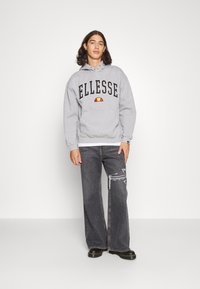 Grijze hoodie met zwarte tekst "ELLESSE" en een rood en geel embleem. Gecombineerd met donkergrijze jeans met een versleten detail. Zwarte schoenen.