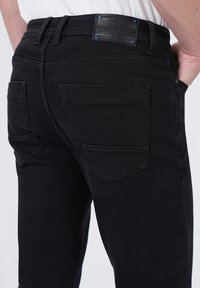 Personne portant un jean slim noir avec des poches arrière et une patch en cuir de marque sur la taille, la main reposant dans la poche avant.