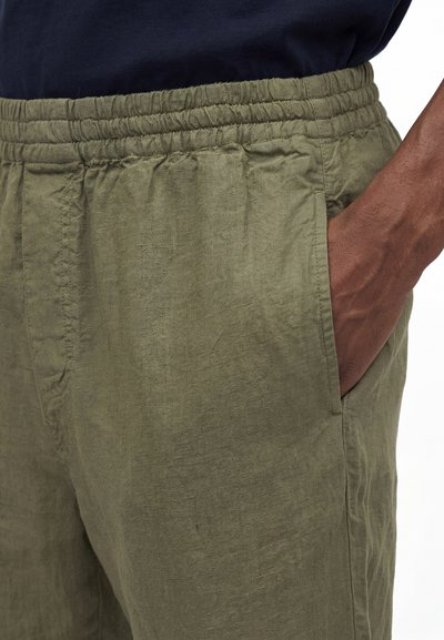 Κοντινό πλάνο ενός ατόμου που φοράει ελαιί πράσινο λινό παντελόνι με ελαστική μέση και το χέρι του να αναπαύεται στην πλαϊνή τσέπη.
