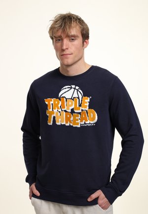 Young man with tousled blond hair wearing a navy sweatshirt with orange and white "Triple Thread" text and basketball graphic.
