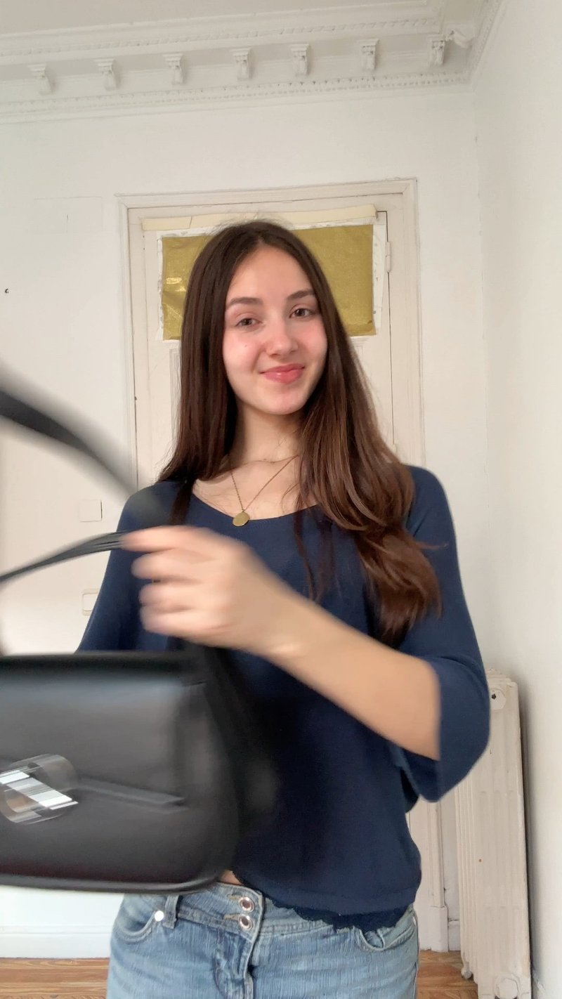 Jeune femme aux longs cheveux bruns portant un haut bleu tient un sac à main noir, souriante, debout à l'intérieur devant une porte blanche.