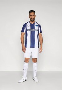 Vīriešu futbola spēlētājs stāv, ģērbies zilā un baltā svītrainā krekla ar "CHECKCARS24" logotipu, baltās šortos, zeķēs un futbola apavos uz vienkrāsaina fona.