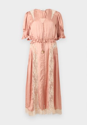 Žena v broskyňovom saténovom midi šatách s čipkovými panelmi, nadýchanými rukávmi, gombíkovými detailmi a čiernymi sandálmi stojaca pred jednoduchým pozadím.