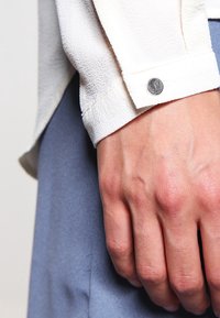 Poignet de chemise blanche texturée avec un bouton-pression métallique, porté sur un tissu bleu clair, présentant une finition lisse et un aspect taillé sur mesure.