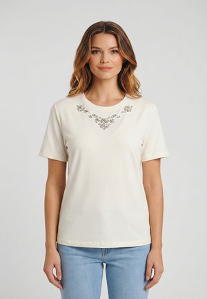 Donna con capelli ondulati fino alle spalle che indossa una maglietta a maniche corte color crema con decorazioni floreali argentate al collo e jeans azzurri.