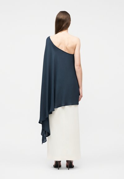 Nő háttal áll, sötétkék egysarkú redőzött felsőt és hosszú fehér szoknyát visel fekete magassarkú szandállal.