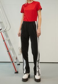 Červené tričko s potiskem, černé tepláky s bílými pruhy a robustní bílé platformové boty. V jedné ruce drží malou bílou tašku.