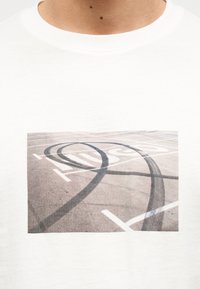 Λευκό μπλουζάκι με τυπωμένη φωτογραφία από σημάδια φρεναρίσματος ελαστικών που σχηματίζουν βρόχους σε έναν άδειο ασφαλτικό χώρο στάθμευσης με λευκές βαμμένες γραμμές.