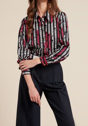 Button-up shirt featuring a bold chain pattern in black, white, and red. Long sleeves with cuffs; paired with high-waisted dark pants.