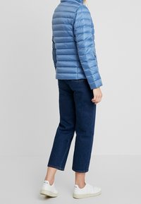 Osoba w jasnoniebieskiej kurtce puchowej, ciemnoniebieskich skróconych dżinsach i białych tenisówkach stojąca na tle jednolitego jasnego tła.