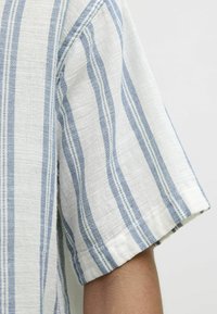 Chemise à manches courtes en tissu texturé. Présente des rayures verticales alternées bleu clair et blanches, avec des détails de poignets repliés.