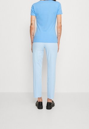 Person trägt ein hellblaues Kurzarmhemd, hellblaue Hose, schwarze Schuhe, steht mit Blick auf eine weiße Wand mit sichtbaren Arm-Tattoos.