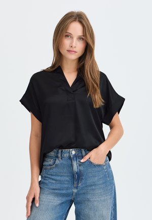 Femme aux longs cheveux châtain clair portant un chemisier noir ample à manches courtes et un jean bleu taille haute, debout avec une main dans la poche.