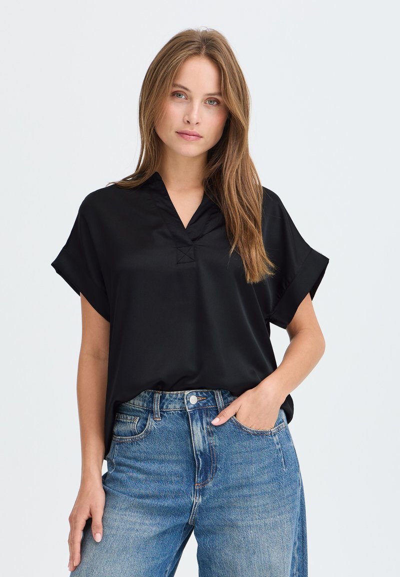 Femme aux longs cheveux châtain clair portant un chemisier noir ample à manches courtes et un jean bleu taille haute, debout avec une main dans la poche.