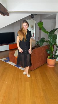 Νέα γυναίκα με μακριά μαλλιά, φορώντας μαύρο φόρεμα με κουμπιά και μπεζ φλατ παπούτσια, στέκεται σε ένα μοντέρνο σαλόνι με πορτοκαλί καναπέ και μεγάλο φυτευμένο φυτό.