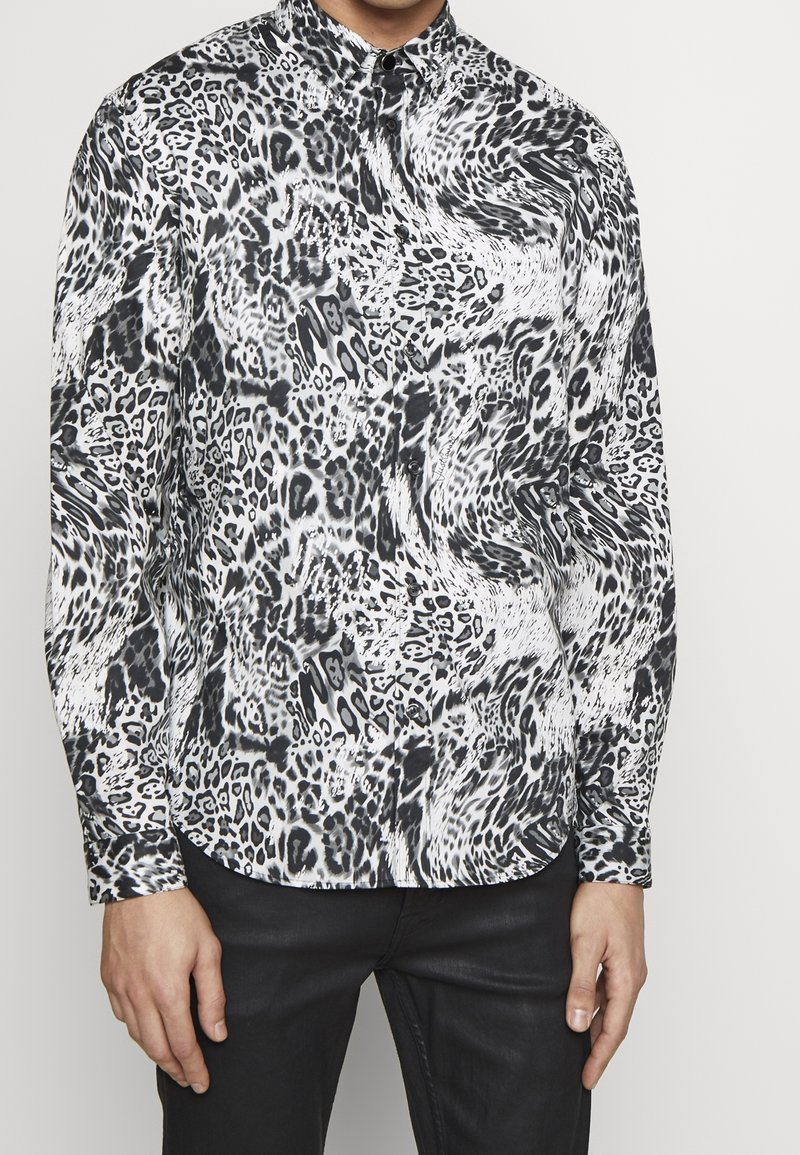 Hombre con camisa de manga larga abotonada con un estampado abstracto de animales en blanco y negro, combinada con pantalones negros.