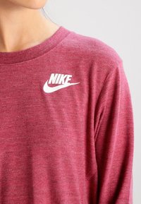 Dlouhý rukáv v barvě vínové od značky Nike, s bílým logem na levé horní straně, vyrobený z měkké, strukturované tkaniny a s kulatým výstřihem.