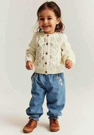 Peutermeisje dat glimlacht, gekleed in een roomkleurig gebreid vest met bloemendetails, losse geborduurde blauwe broek en bruine veterschoenen.