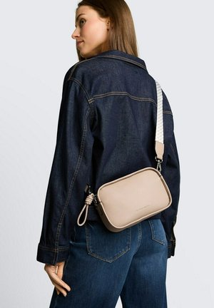 Vrouw draagt een donker denimjack en spijkerbroek, met een beige schoudertas met een witte band over haar schouder.