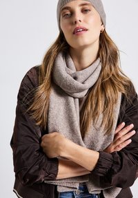 Écharpe beige en tissu tricoté doux, avec une texture côtelée et un design superposé et lâche, associée à une veste marron foncé avec des manches bouffantes.
