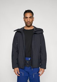 Casaco impermeável azul-marinho com fecho de correr frontal, capuz ajustável e bolsos. Usado sobre uma camisa preta e calças azuis com bolsos e botões.