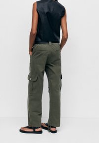 Veste en cuir noire sans manches, pantalon cargo vert olive avec poches latérales, et sandales plates noires, vus de dos sur un fond uni.