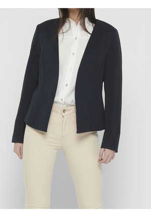 Femme portant un blazer noir sur mesure par-dessus une chemise blanche à boutons et un pantalon beige, debout devant un fond gris clair.