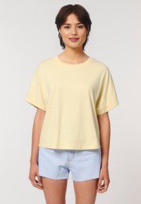 Young woman smiling, wearing a loose-fitting yellow t-shirt and light blue shorts, against a plain background.