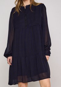 Mörkblå, långärmad klänning med rund halsringning, knäppning fram och en lager-på-lager kjol. Har ett subtilt geometriskt mönster i ljusare blått.