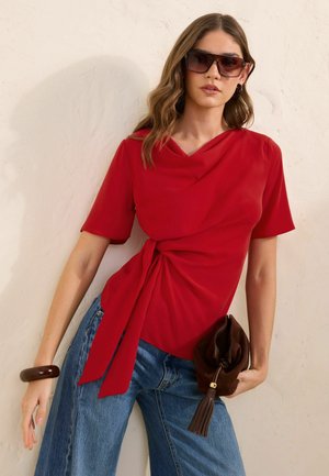 Vrouw met oversized bruine zonnebril, rode korte mouw wikkel top gestrikt in de taille, blauwe jeans, houdt een bruine clutch met kwastje en een bruine armband vast.