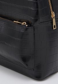 Czarna torba ze skóry wytłaczanej w wzór krokodyla, z złotym zamkiem błyskawicznym, gładką teksturą i wyraźnie zarysowanym kształtem, posiadająca przednią kieszeń.