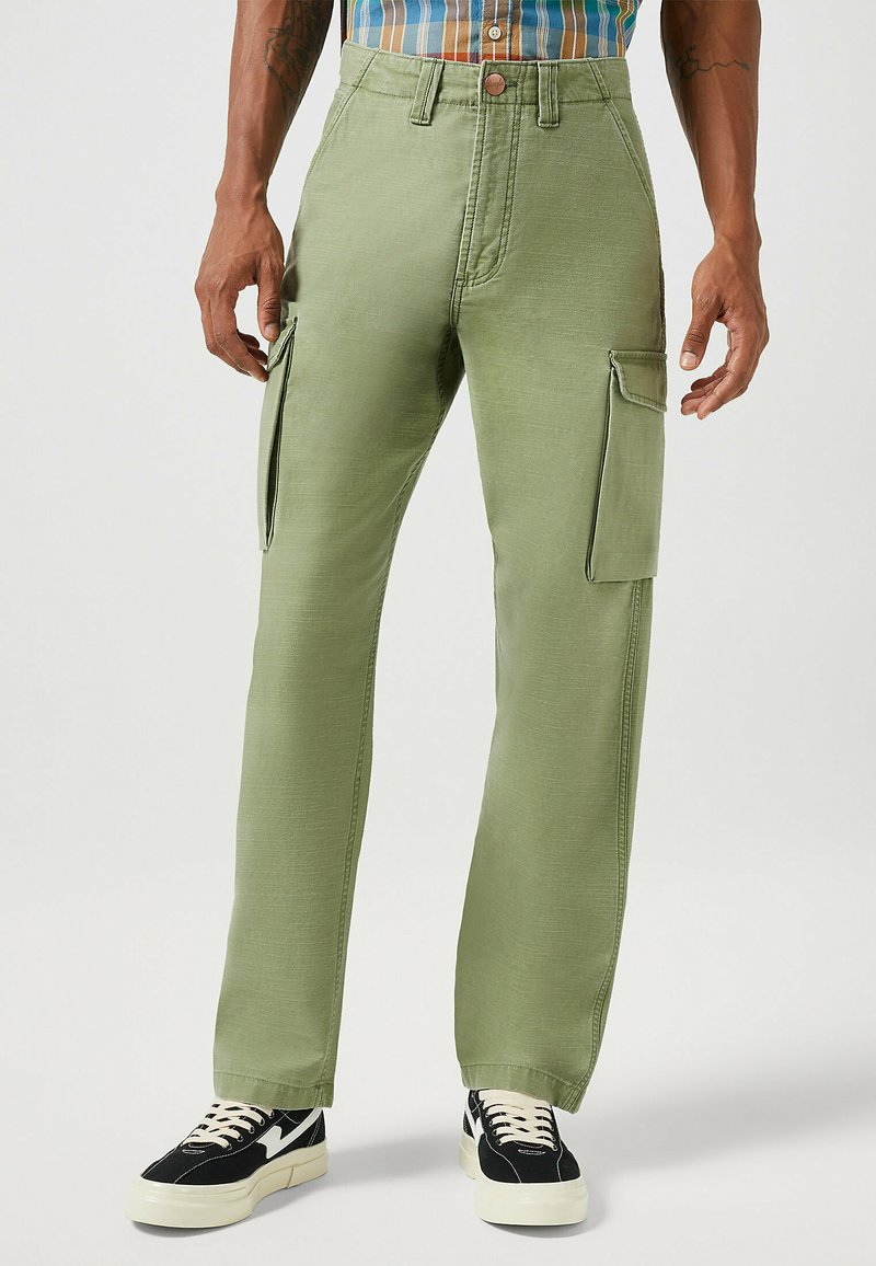Pantalones cargo verdes con un corte recto, con bolsillos laterales y cierre de botón. Combinados con zapatillas negras y blancas. Textura de tela suave.