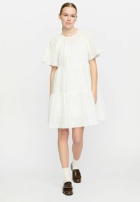 Hvid kjole med rund halsudskæring, korte fl�æseærmer, lagdelt design og øjelet detaljer. Sammen med brune loafers og hvide sokker.