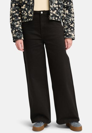 Jean noir à taille haute et jambes larges avec une texture subtile, associé à une veste à motifs floraux. Chaussures argentées avec semelles en caoutchouc.