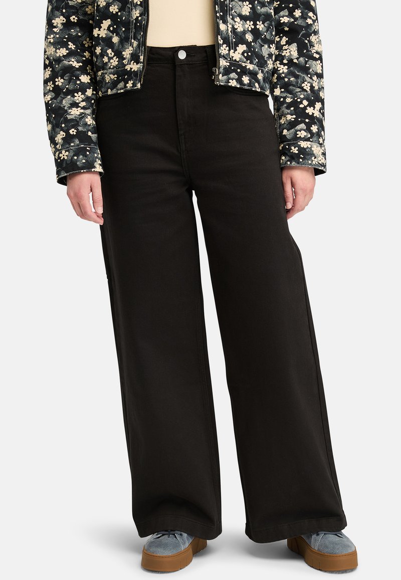 Vaqueros negros de pierna ancha con cintura alta y textura sutil, combinados con una chaqueta de estampado floral. Zapatos en color plata con suelas de goma.