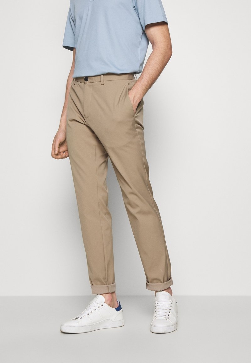 Pantalones chinos beige de corte recto, con cierre de botón, bolsillos laterales y bajos remangados, combinados con zapatillas blancas y una camisa azul clara.