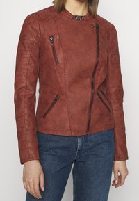 Kobieta ubrana w rdzawą skórzaną kurtkę motocyklową z asymetrycznym zamkiem błyskawicznym i pikowanymi detalami, w połączeniu z niebieskimi dżinsami.