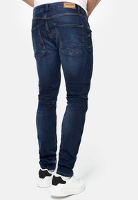 Jeans de denim azul oscuro con ajuste slim, con bolsillos traseros y una etiqueta de cuero marrón clara con marca en la cintura. Combinados con zapatillas blancas.