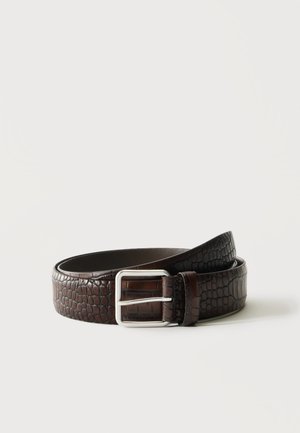 Ceinture en cuir marron foncé à texture crocodile, boucle en métal argenté, enroulée sur fond blanc uni.