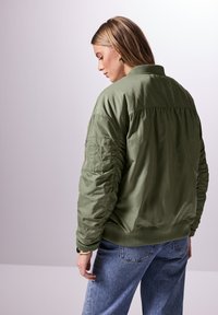 Vihreä bombere-takki, jossa on resoritettu kaulus ja hihansuut sekä vasemmassa hihassa paikka-tasku ja sileä, kiiltävä pinta.