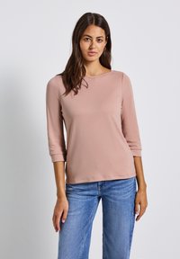 Frau mit langen dunklen Haaren, die ein hellrosa Oberteil mit drei Viertelärmeln und eine blaue Jeans trägt, steht vor einem schlichten, hellen Hintergrund.