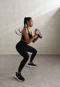 Vêtements de sport noirs avec un débardeur ajusté et des leggings, tenant une plaque de poids, effectuant un squat sur un sol lisse et gris clair.