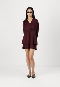 Bordo apvyniota suknelė su ilgomis rankovėmis, pakopinėmis volanėlėmis apatinėje dalyje ir prigludusia juosmens dalimi. Modelis dėvi juodas smailias basutes ir saulės akinius.
