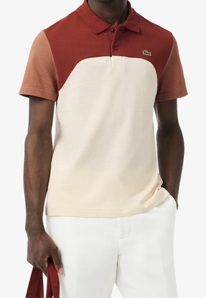 Homme portant un polo à manches courtes avec des blocs de couleur rouille et beige, petit logo crocodile, pantalon blanc et tenant une bandoulière de sac rouge.