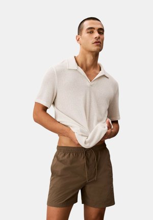 Uomo che sistema una camicia a maniche corte color crema con una trama, indossa pantaloncini marroni con coulisse su uno sfondo chiaro e uniforme.
