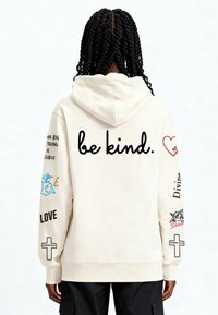 Kremowa bluza z kapturem ze ściągaczem, z napisem i kolorowymi grafikami na plecach oraz na obu rękawach, w tym krzyżem i kwiatowymi wzorami.