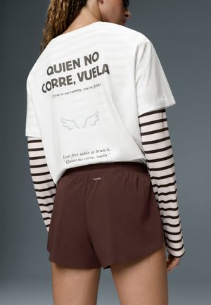 Personne aux cheveux tressés portant des lunettes de soleil, un t-shirt blanc avec un texte en espagnol et en anglais, des manches longues rayées et un short marron sur fond gris.