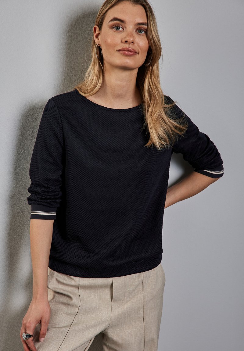 Dunkelblaues Sweatshirt mit Textur und langen Ärmeln, Rundhalsausschnitt und gestreiften Akzenten an den Bündchen, kombiniert mit hellbeigen Hosen.