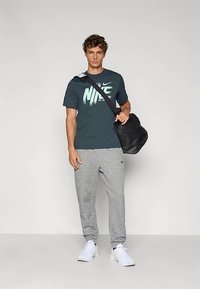 Camiseta azul oscuro de Nike con un logo claro, pantalones deportivos grises y zapatillas blancas. Bolsa negra colgada al hombro. Fondo liso.